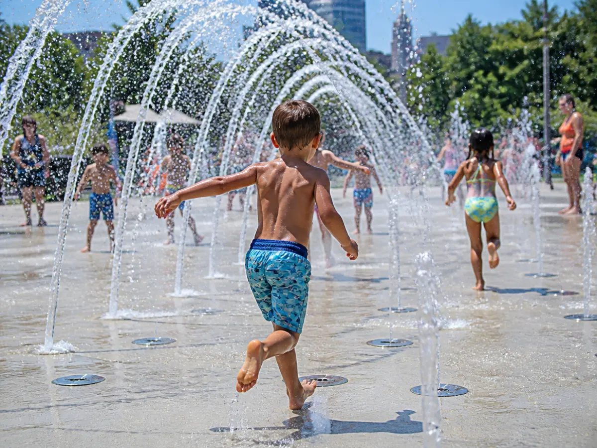 Illustration: children running through ground water jets and spray arches at a splash pad in a city park on a hot summer day